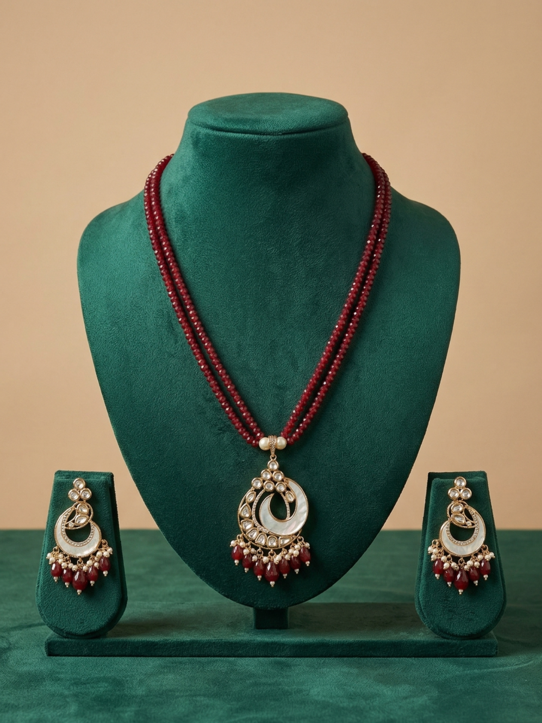 A traditional Indian jewelry set featuring a long red beaded necklace with a silver and white crescent mop pendant, accompanied by matching earrings, displayed on a green velvet mannequin bust.