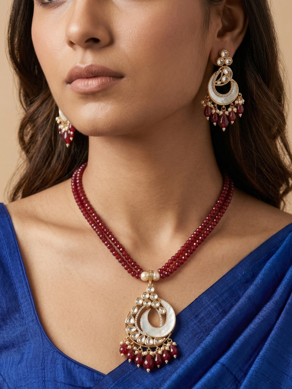 A close-up of a woman wearing a traditional Indian jewelry set featuring a beaded red necklace with a large gold and white crescent mother of pearl pendant, matching drop earrings, and a royal blue saree.
