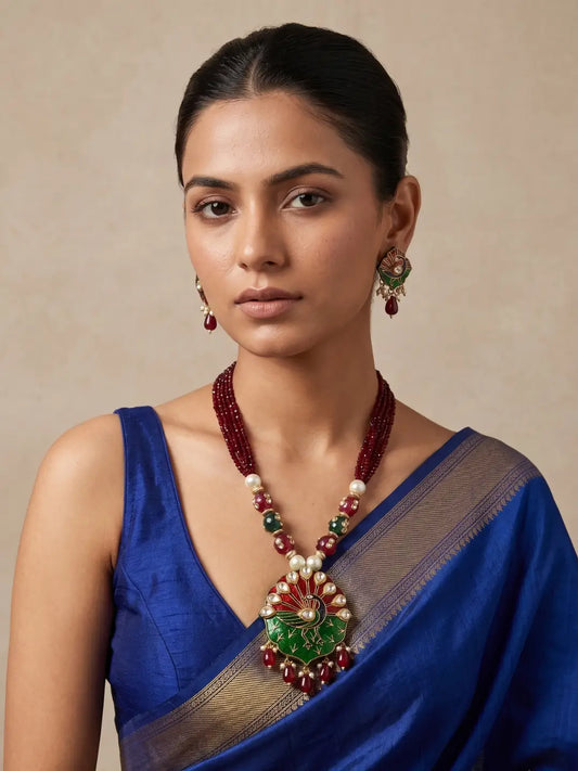 A woman is wearing a royal blue saree with a gold border and a traditional Indian statement necklace and matching earrings. The necklace features a large green and red floral pendant with white peacock detailing and pearl-like accents, and the earrings have red and green details.