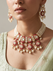 A close-up of a model wearing an ornate Indian-style jewelry set featuring a red and gold necklace with pearl-like white beads, matching drop earrings, and a light green embroidered blouse.