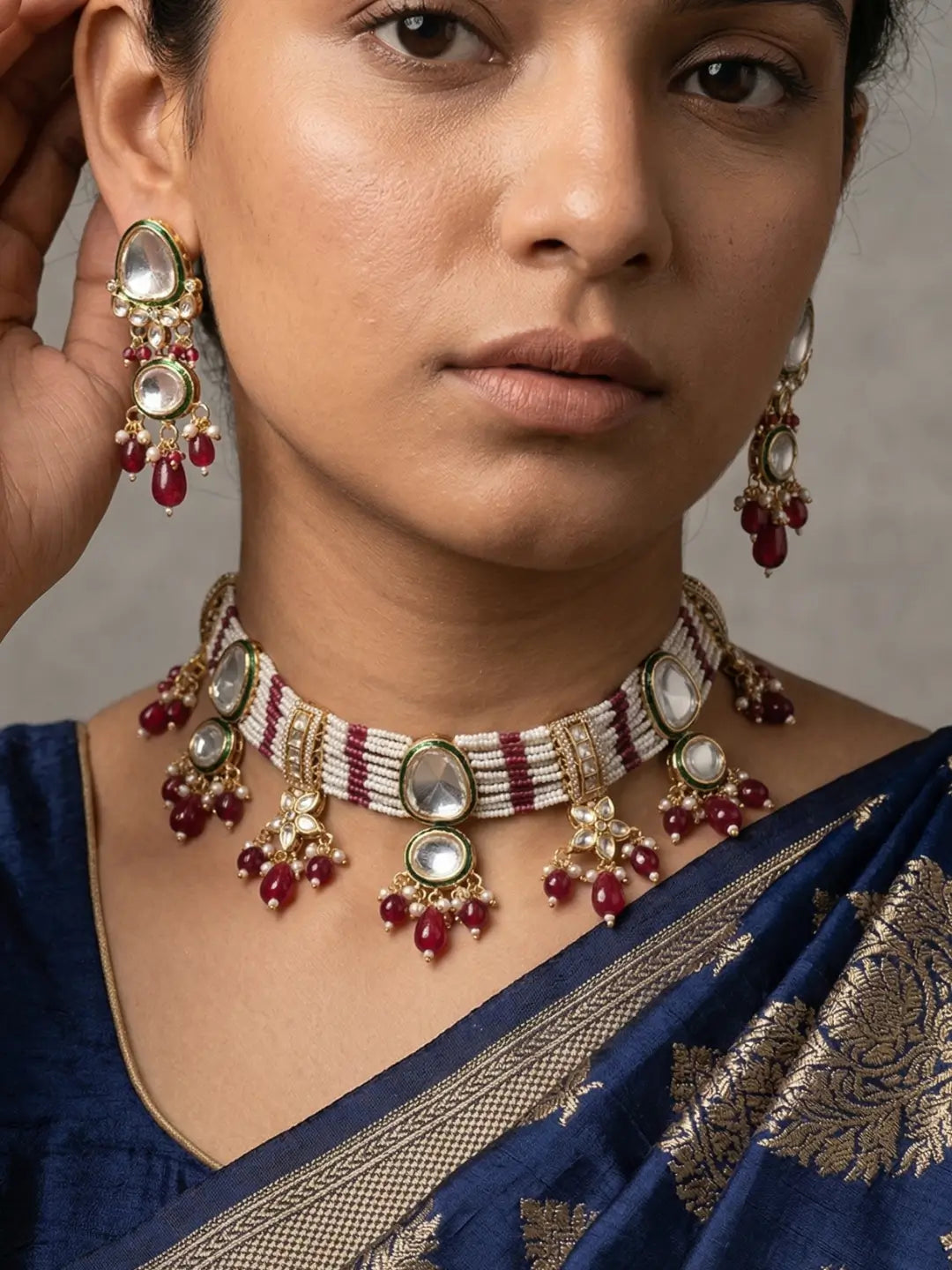 A close-up of a woman wearing a traditional Indian gold-plated red choker necklace and matching drop earrings with white stones and red beads, paired with a navy blue saree.