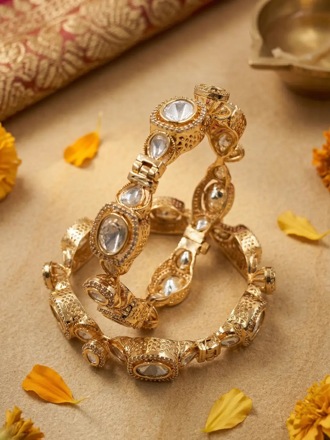 A pair of ornate gold-toned bangles decorated with clear stones, arranged on a beige surface with scattered yellow flower petals and a rolled patterned fabric in the background.