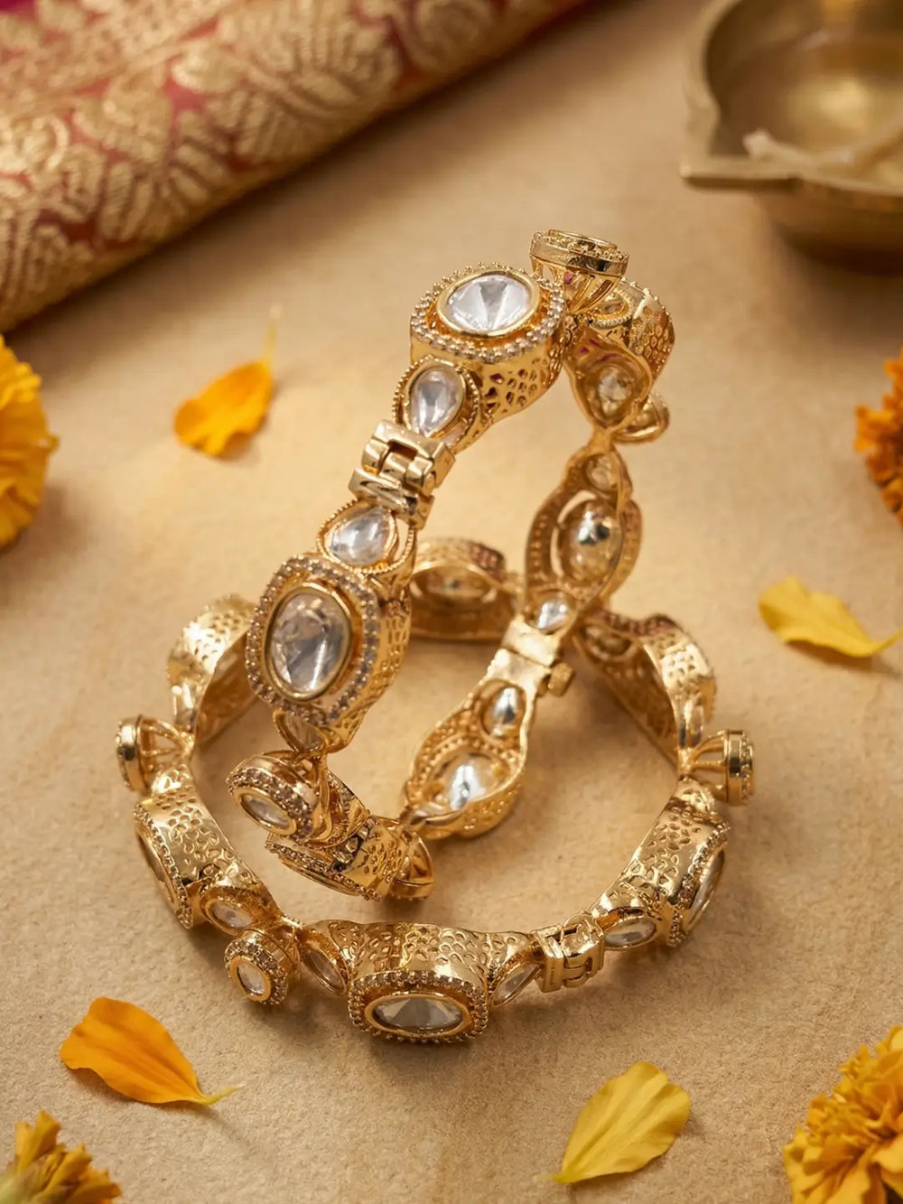A pair of ornate gold-toned bangles decorated with clear stones, arranged on a beige surface with scattered yellow flower petals and a rolled patterned fabric in the background.