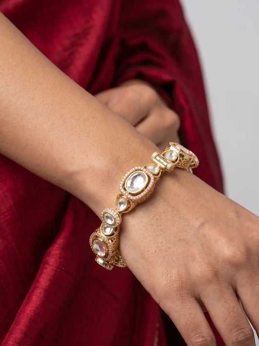A rose gold-toned traditional Indian kada bracelet with embedded white polki stones, shown worn on a person's wrist against a deep red fabric background.
