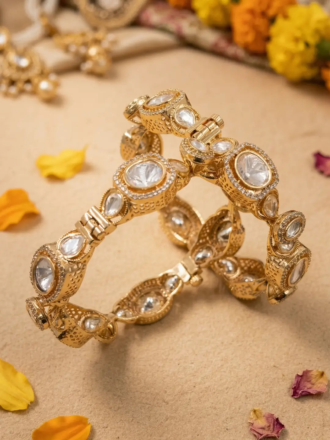 A set of rose gold-plated traditional Indian Kundan bangles adorned with clear polki crystals, arranged on a beige textured surface with scattered yellow and pink flower petals.