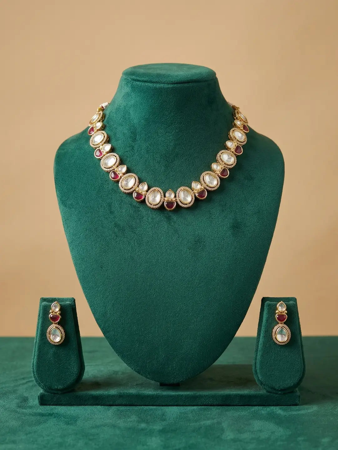 Gold-toned jewelry set displayed on a dark green velvet mannequin bust, featuring a necklace with round white stones and red gemstone accents, plus matching drop earrings on small standees below.