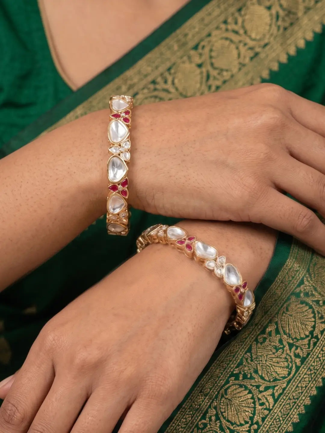 Gold-toned bangles studded with white teardrop-shaped polki kundan stones and small red gemstone accents, shown worn on a person's wrist and hand over a green embroidered sari.