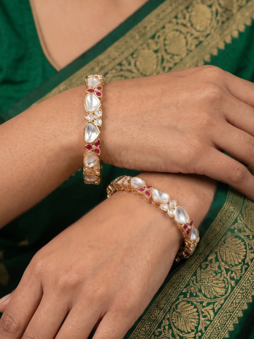 Gold-toned bangles studded with white teardrop-shaped polki kundan stones and small red gemstone accents, shown worn on a person's wrist and hand over a green embroidered sari.