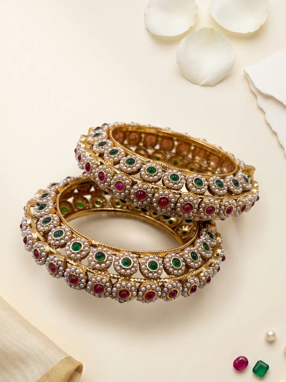 A pair of ornate gold-plated ethnic kada bangles decorated with rows of white stones, green emeralds, and red rubies, arranged on a light cream background with decorative white petals, perfect for wedding, bridal and festive wear 