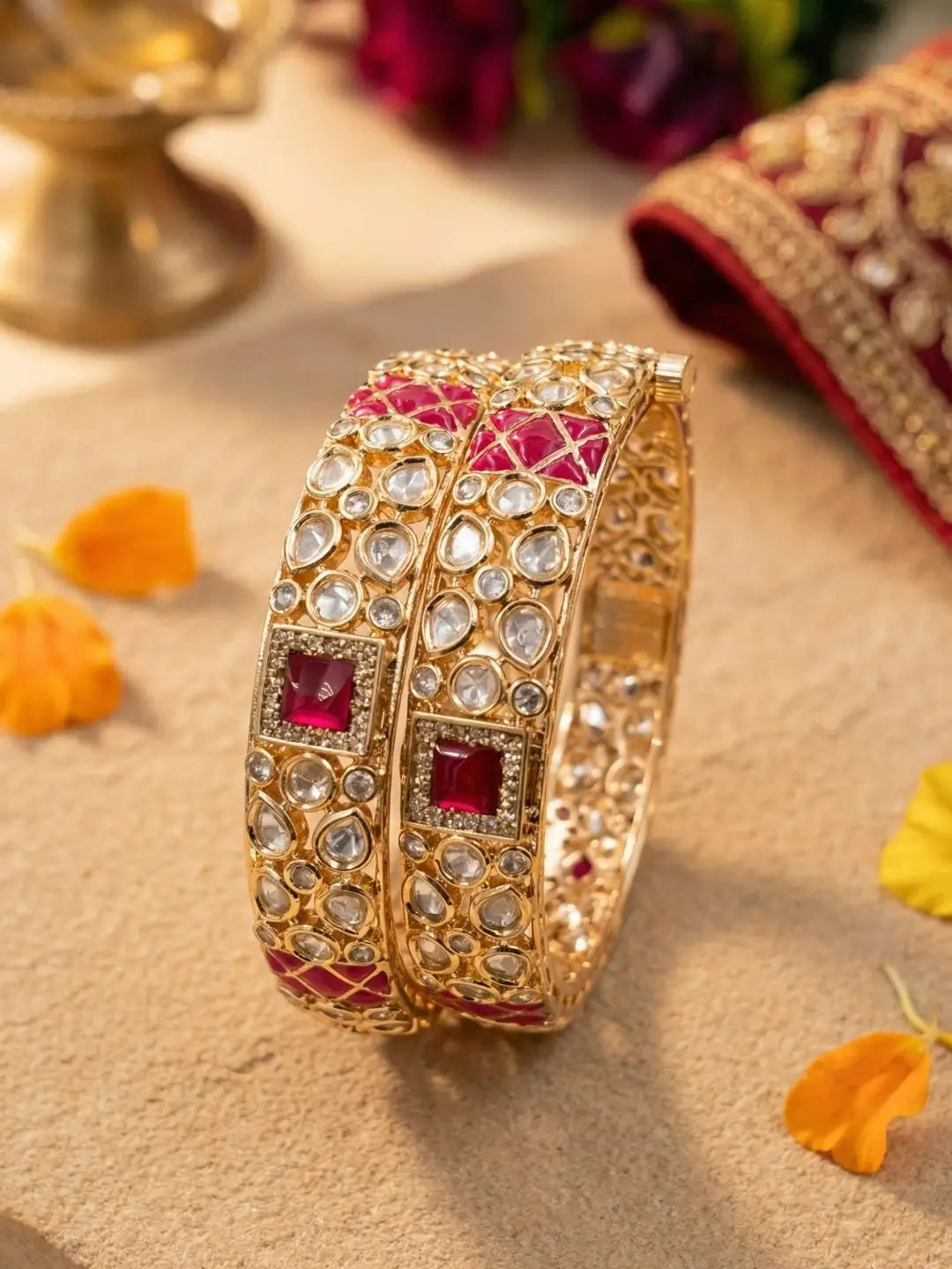 A pair of gold-plated Indian bangles decorated with clear crystals and red enameled inlays, displayed on a beige surface with decorative flower petals and traditional background elements.