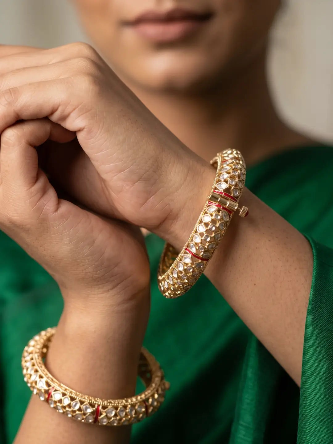 A close-up of a person wearing two ornate gold-plated uncut polki-style bangles with red and white stone embellishments, set against a soft-focus background and a green garment.