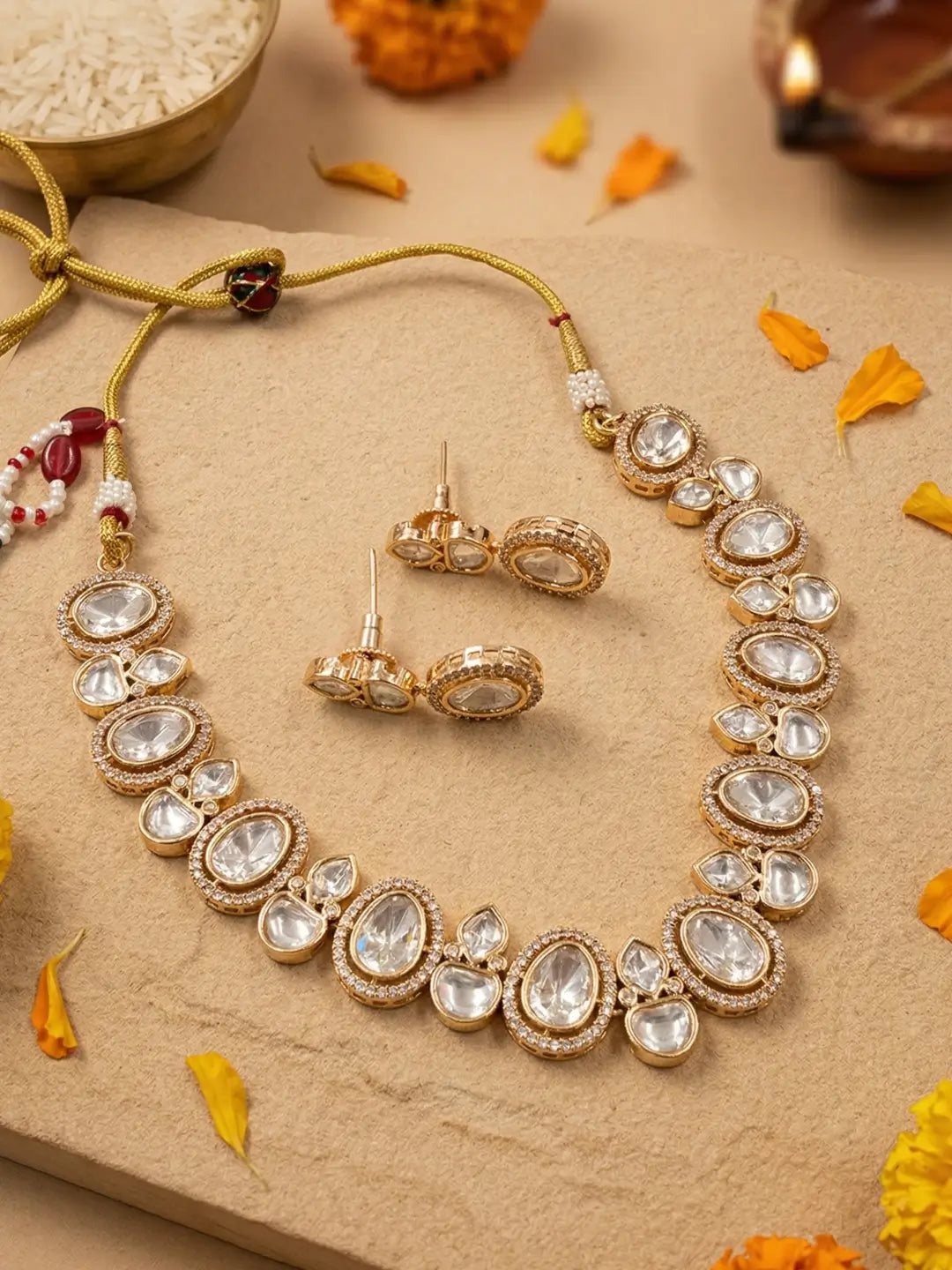 Gold-toned traditional Indian jewelry set featuring a necklace and matching earrings adorned with clear teardrop-shaped stones, arranged on a beige stone surface with scattered yellow flower petals.