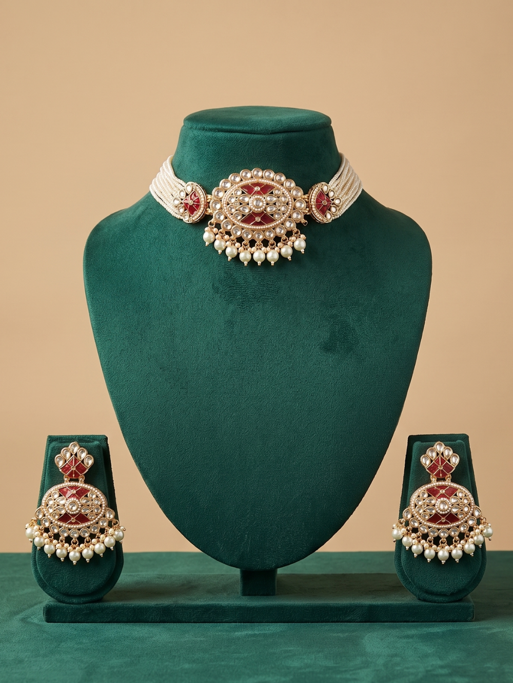 A traditional Indian ruby-red kundan-and-pearl beaded choker jewelry set displayed on a dark teal velvet mannequin bust. The set includes a gold-plated choker necklace with green bead fringe and matching drop earrings, set against a solid beige background.