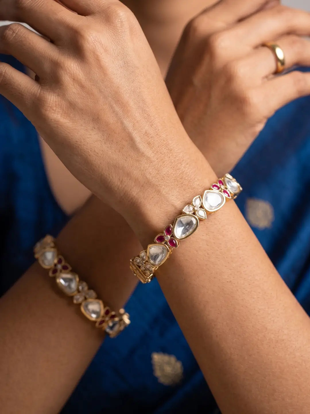 A close-up of a person's hands and arms wearing two gold-plated kundan bangles adorned with white stones and small red gem accents, paired with a blue traditional outfit.