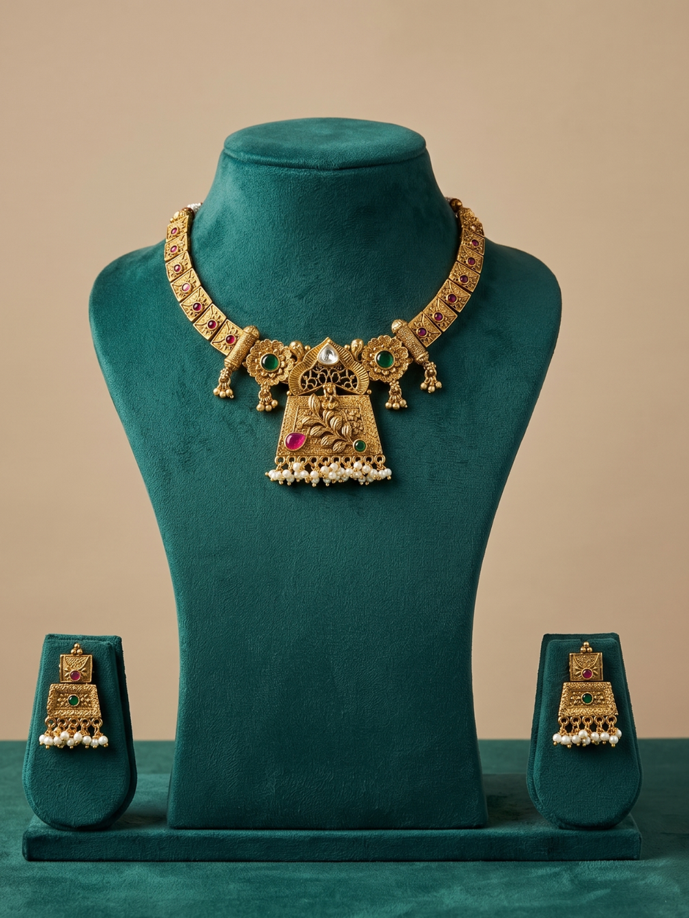 An ornate gold-toned traditional necklace with matching drop earrings displayed on a teal velvet jewelry bust set against a warm beige background.