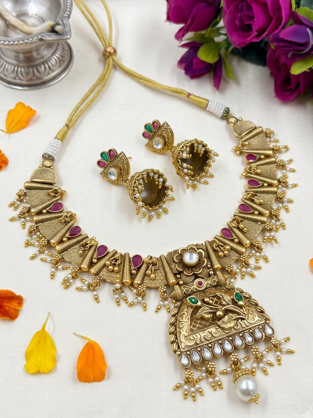 Gold-toned traditional Indian antique jewelry set featuring a statement necklace, matching earrings, and a small pendant, arranged on a light surface with decorative flower petals and flowers.