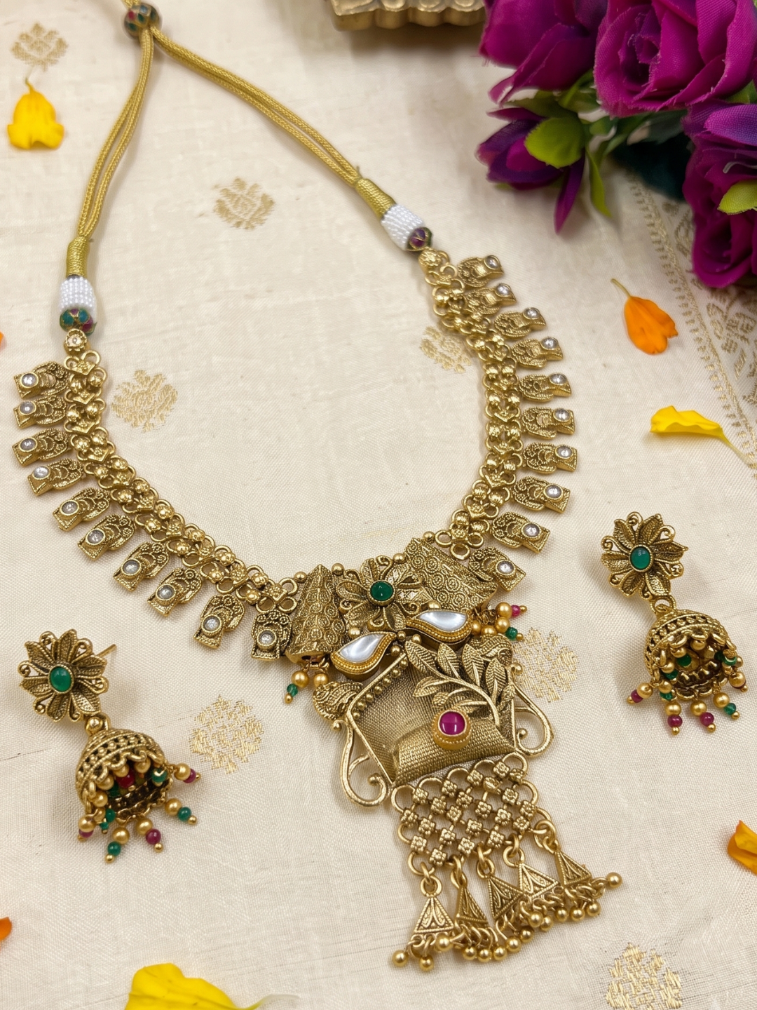 An ornate gold-toned Indian antique gold jewelry set featuring a necklace, matching earrings, and a pendant, decorated with faux pearls, green and red stones, displayed on a cream fabric background with decorative flowers.