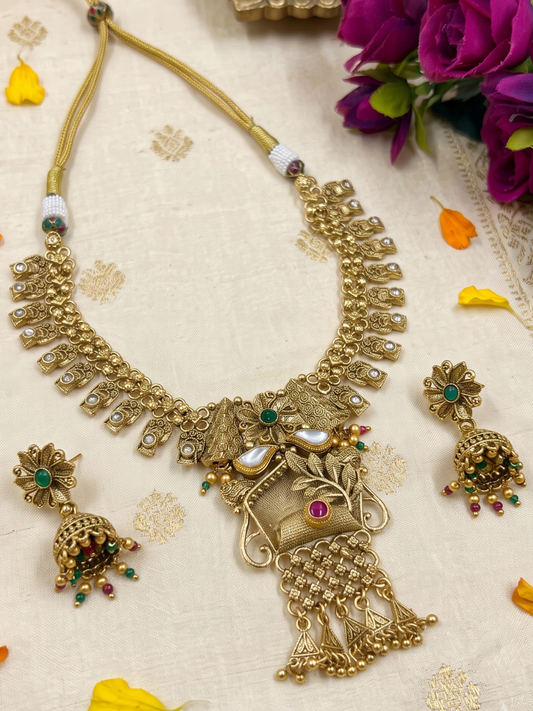 An ornate gold-toned Indian antique gold jewelry set featuring a necklace, matching earrings, and a pendant, decorated with faux pearls, green and red stones, displayed on a cream fabric background with decorative flowers.