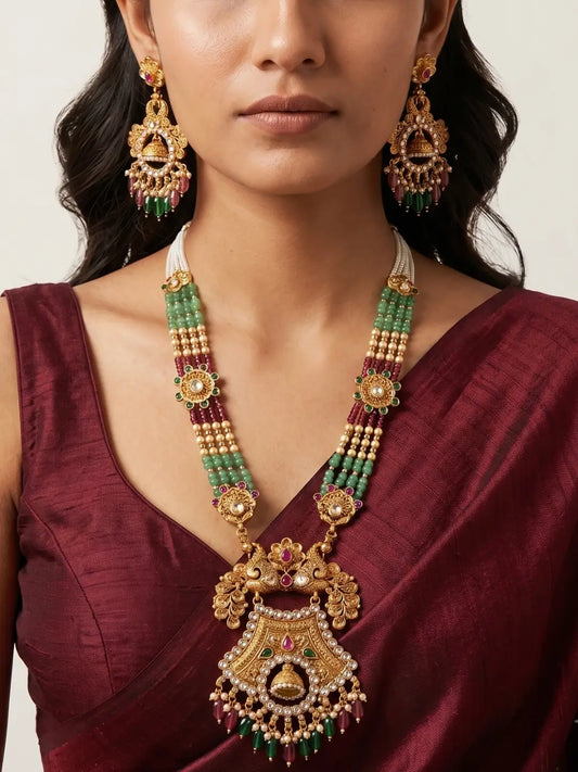 A close-up of a model wearing an ornate, traditional antique-gold necklace with green and red bead strands, matching earrings, and a deep red saree, set against a light, neutral background.