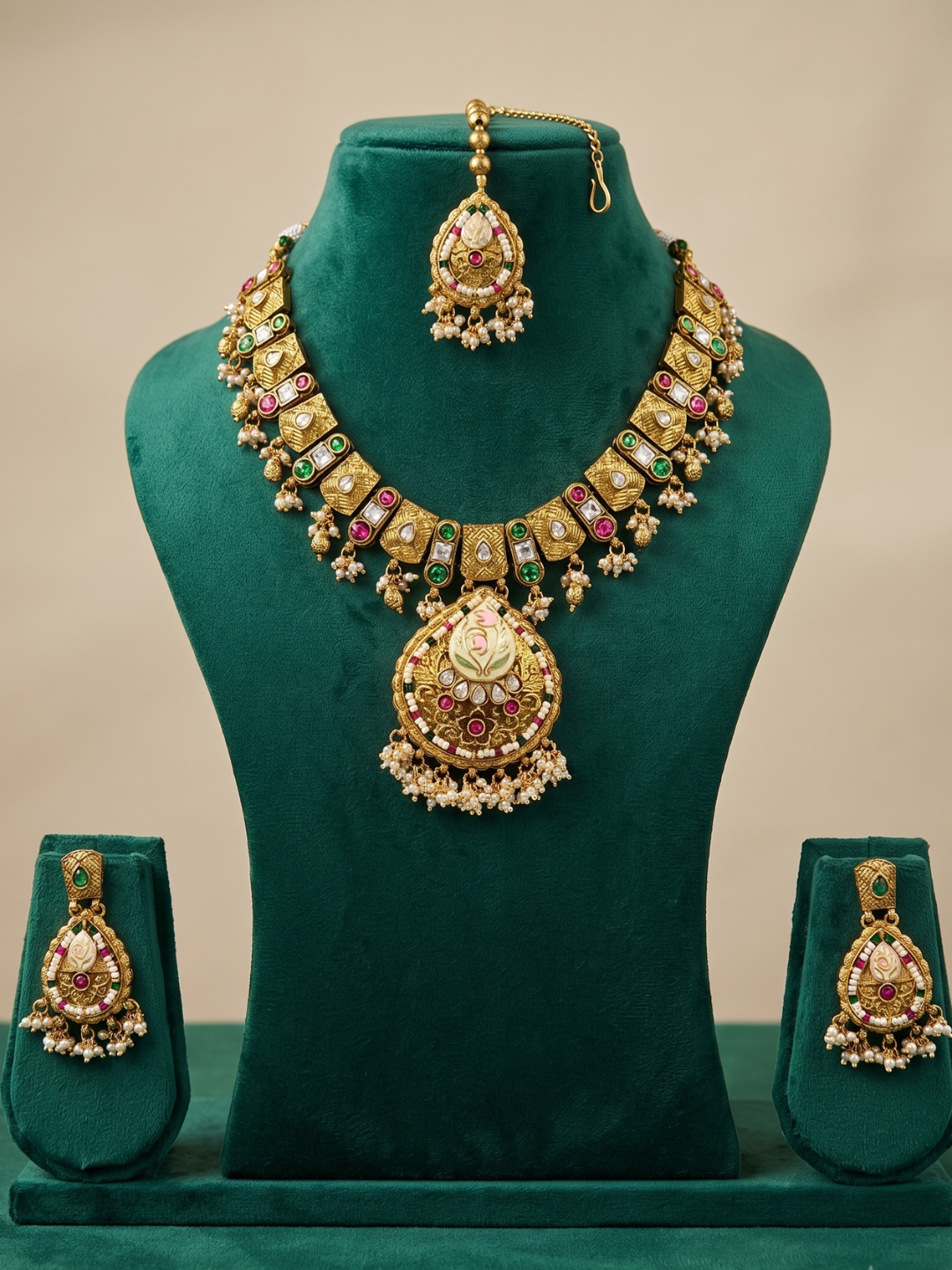 An ornate Indian gold jewelry set displayed on a dark green velvet mannequin bust, including a necklace with a large pendant, a matching maang tikka, and two matching earrings.