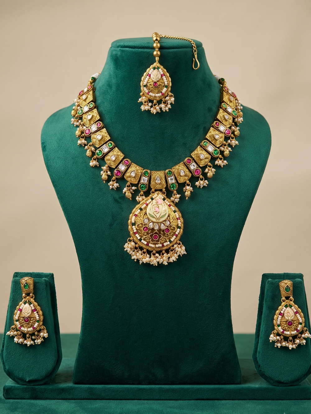 An ornate Indian gold jewelry set displayed on a dark green velvet mannequin bust, including a necklace with a large pendant, a matching maang tikka, and two matching earrings.