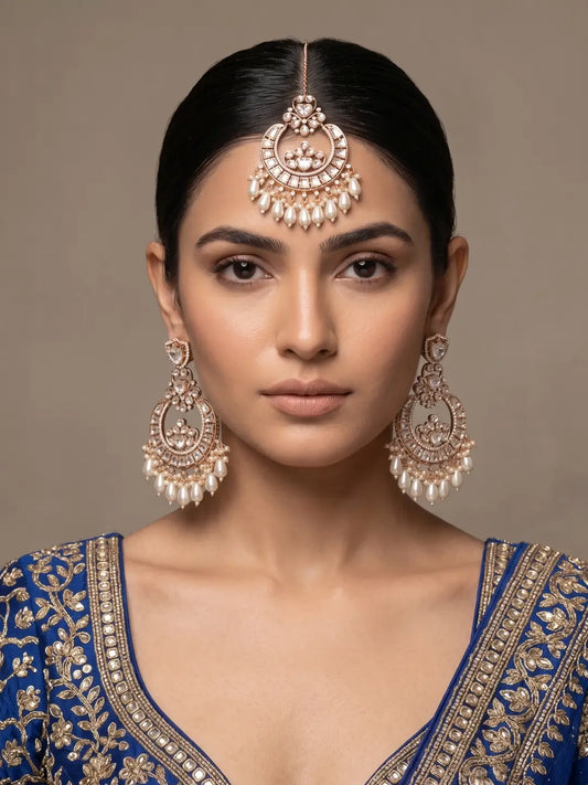 A woman wearing traditional Indian jewelry, featuring a gold-plated oversized kundan maang tikka with white stones and matching chandbali earrings, paired with a blue embroidered blouse.