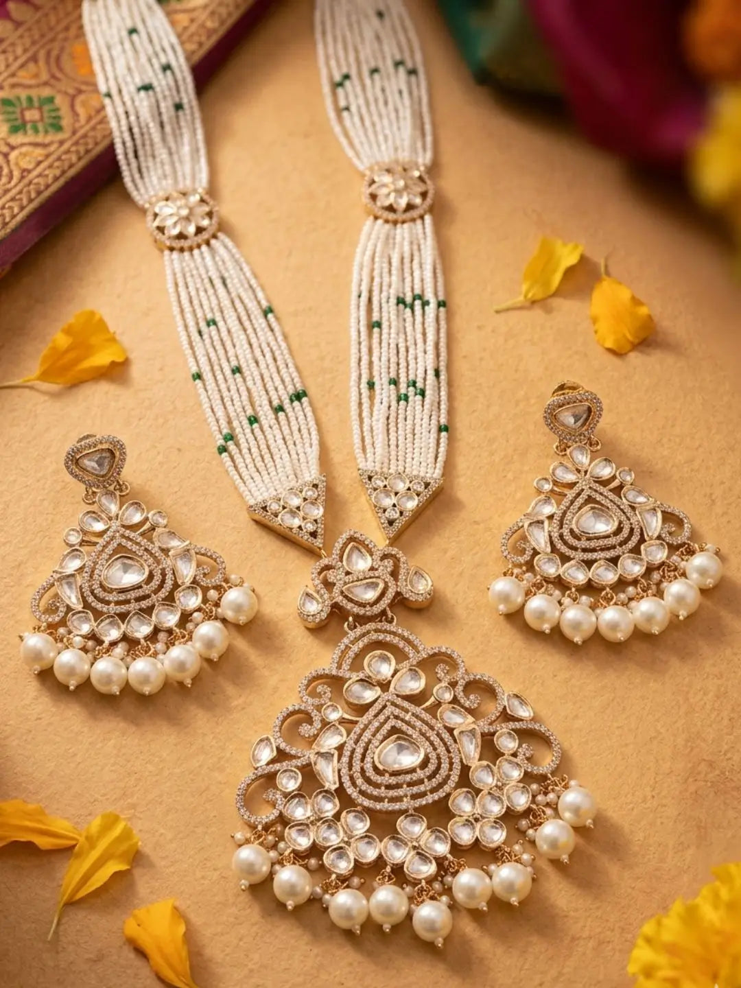 A traditional Indian bridal jewelry set featuring a gold-plated necklace and matching earrings adorned with white pearls, clear stones, and green bead accents, arranged on a warm tan background with scattered yellow flower petals.