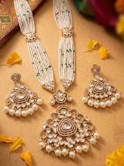 A traditional Indian bridal jewelry set featuring a gold-plated necklace and matching earrings adorned with white pearls, clear stones, and green bead accents, arranged on a warm tan background with scattered yellow flower petals.