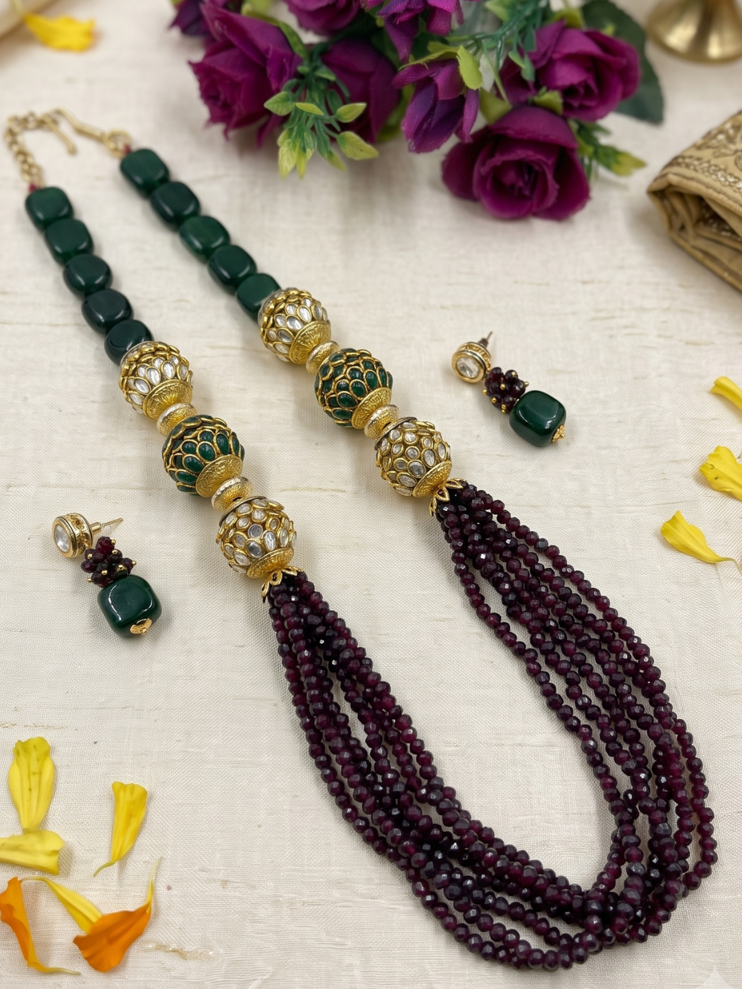 A traditional Indian beaded jewelry set featuring a necklace and matching earrings. The necklace has dark green square beads, ornate kundan and green patterned pacchi beads, and cascading multi-strand dark maroon beaded tassels.
