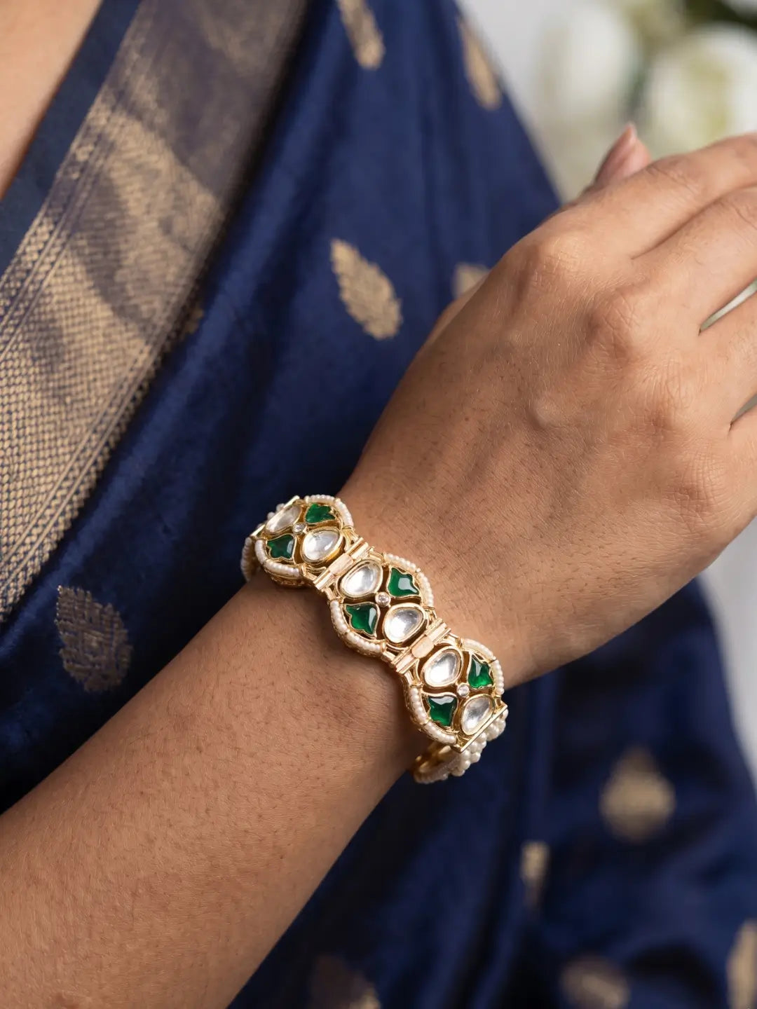 A close-up of a person's wrist wearing an ornate gold-toned traditional Indian bracelet decorated with green meena and polki stones paired with a navy blue saree.