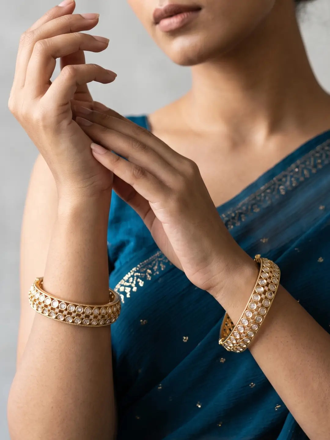 A close-up of a woman wearing ornate gold bangles adorned with small white stones, paired with a teal blue sari. She is holding her hands together in front of her chest, showcasing the jewelry against a soft gray background.