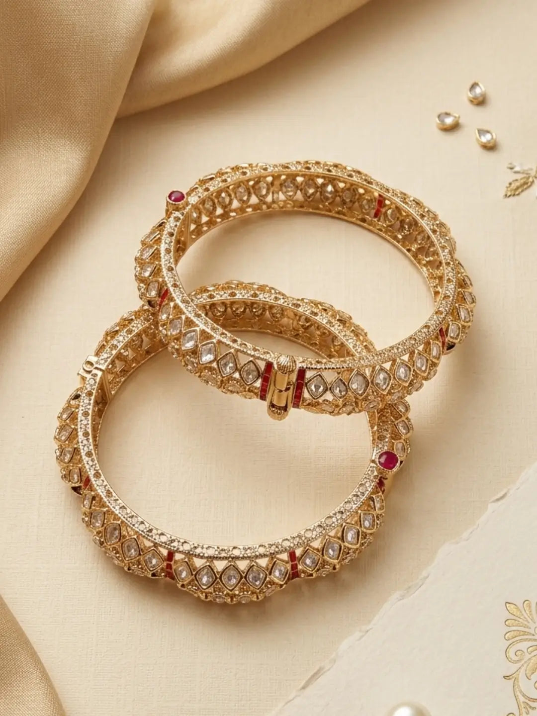 Gold-plated Indian red kundan polki style bangles adorned with white stones and small red gem accents, arranged on a beige satin fabric background.