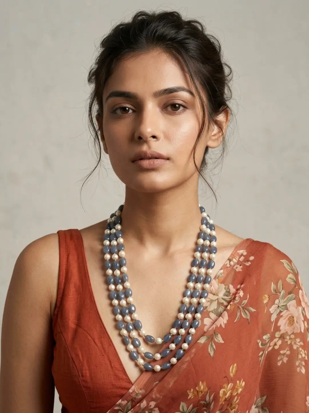 A woman wearing a multi-strand beaded necklace with blue-gray and cream beads, paired with a rust-orange floral saree, set against a neutral textured background.