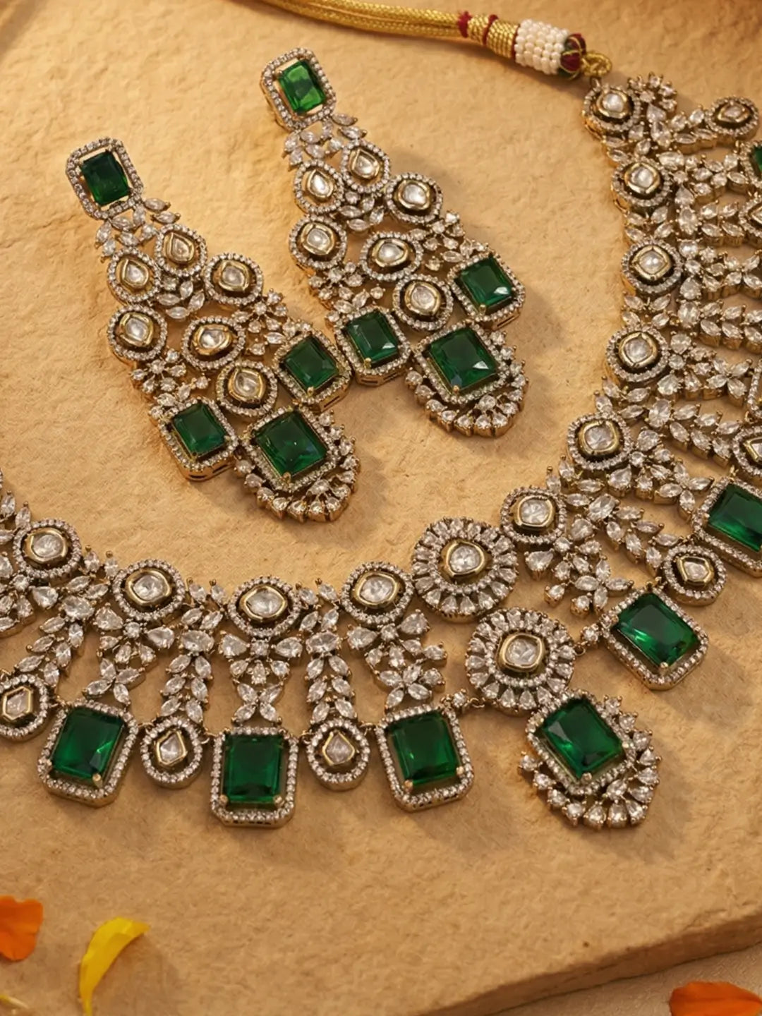 An ornate Indian ethnic jewelry set featuring a silver-tone necklace and matching earrings embellished with green rectangular emerald stones and round polki stones, displayed on a textured beige stone surface.